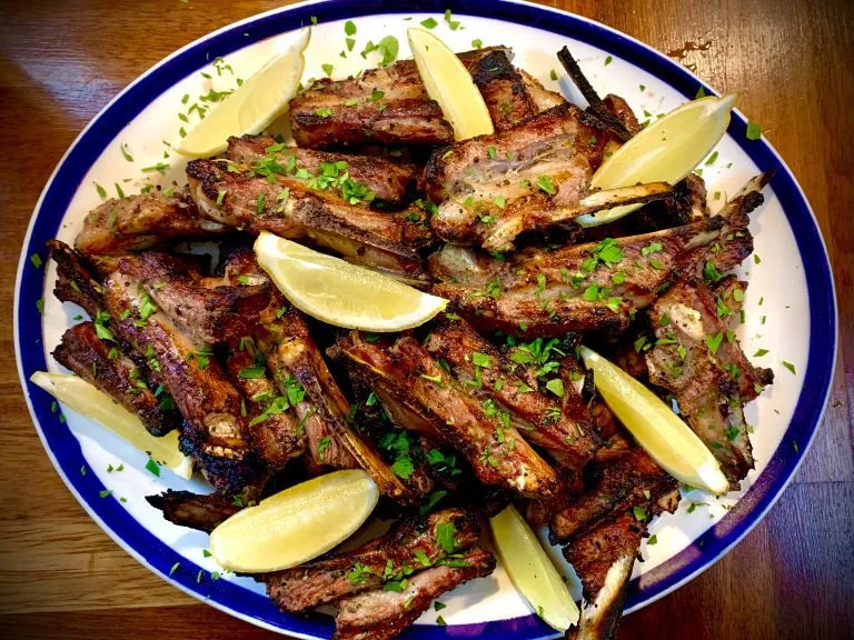 Grilled Greek lamb ribs on a plate with lemon wedges and chopped parsley