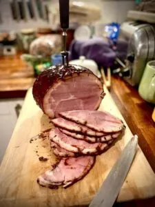 Whole glazed ham with a stack of pink slices, a carving knife and fork, on a board