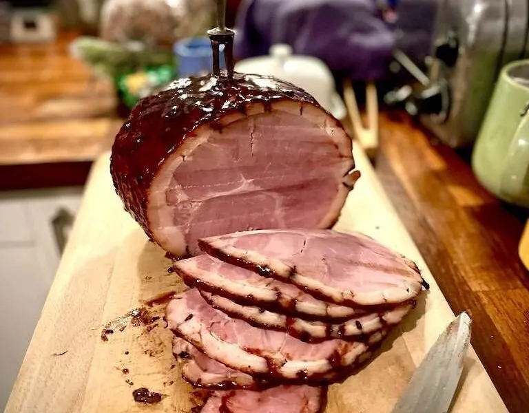 Whole glazed ham with a stack of pink slices, a carving knife and fork, on a board